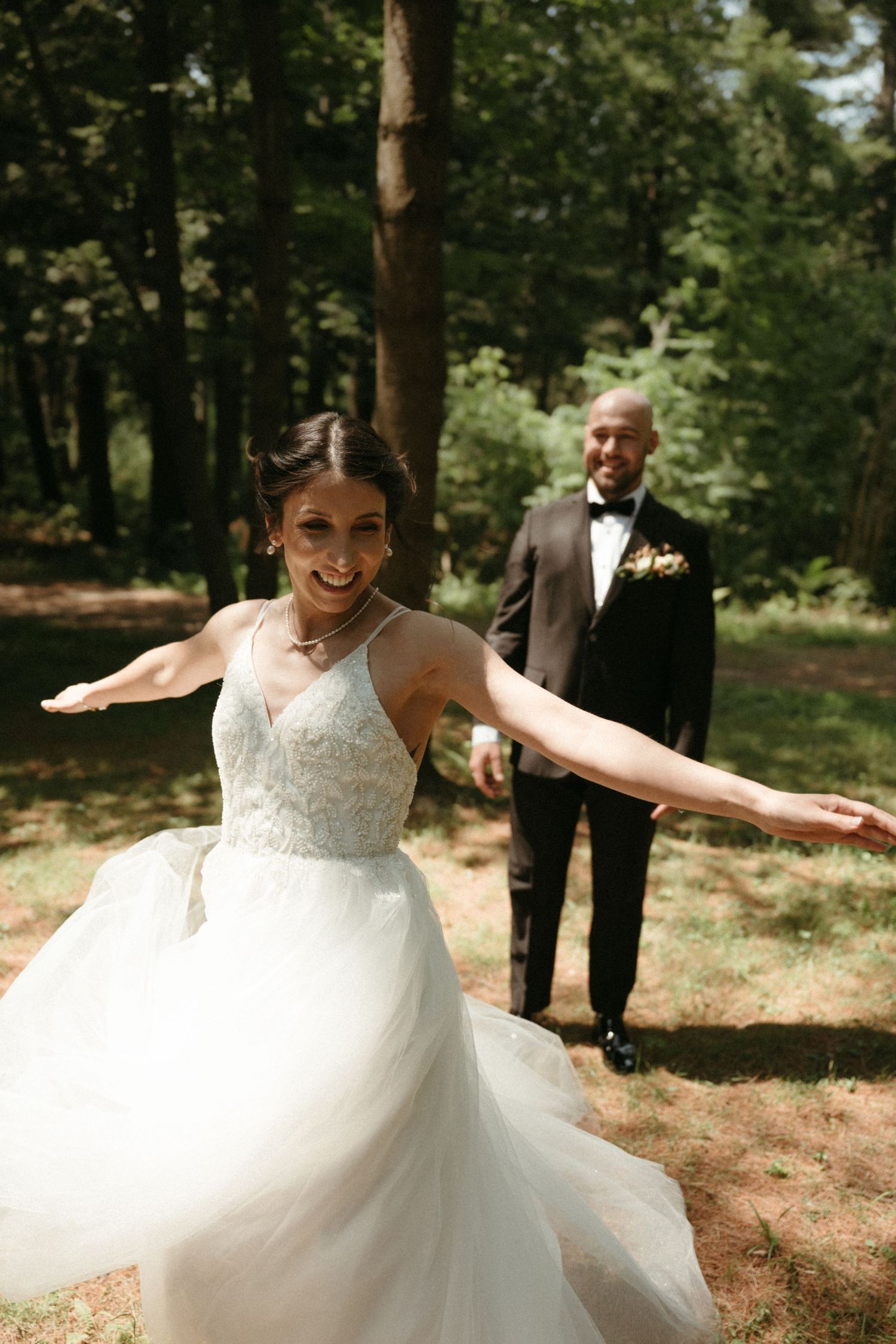 Bride in white lace gown twirling outdoors with groom in black tuxedo standing behind her in a forest setting.