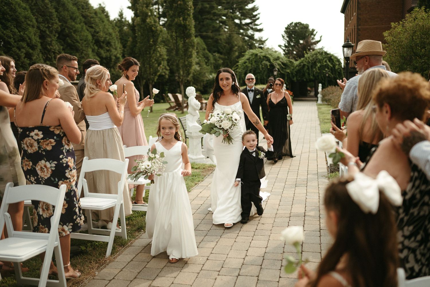 Bride walks down outdoor aisle with flower girl and ring bearer as guests celebrate at a garden wedding.