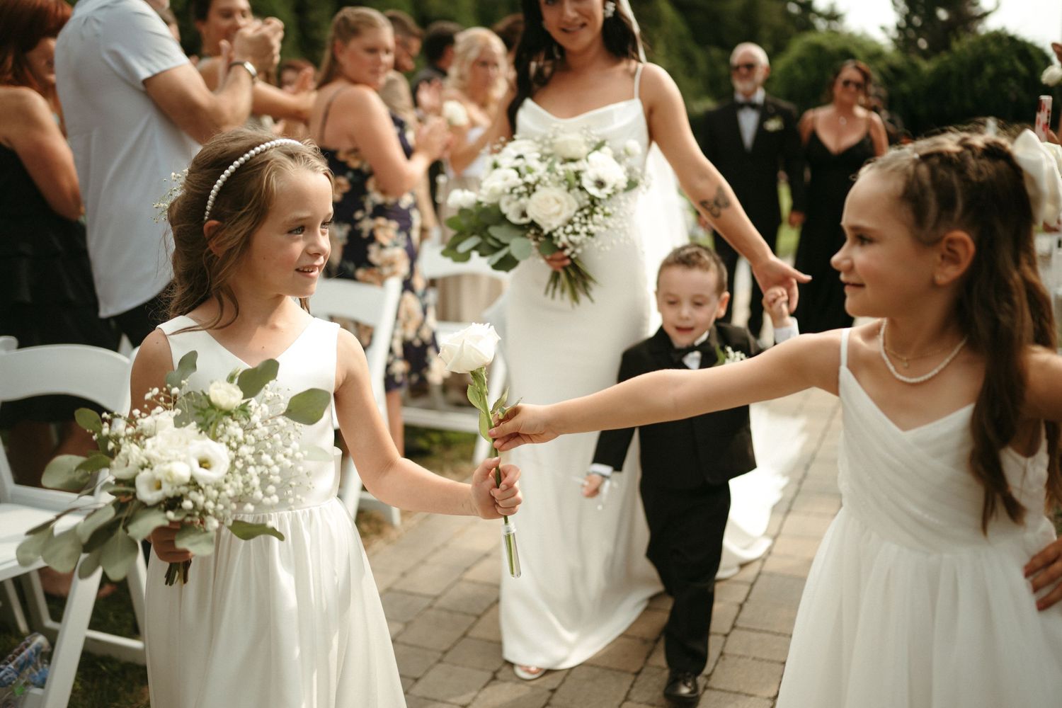 Two flower girls in white dresses holding bouquets smile during outdoor wedding ceremony procession.