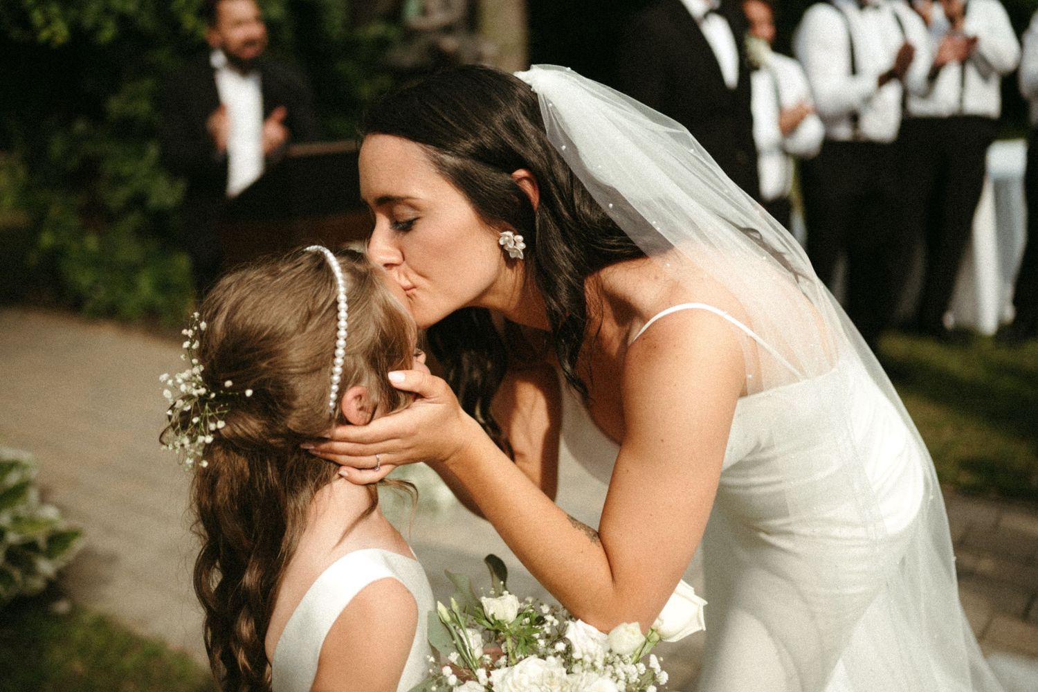 Bride in white gown and veil kisses a flower girl wearing a floral crown at an outdoor wedding.