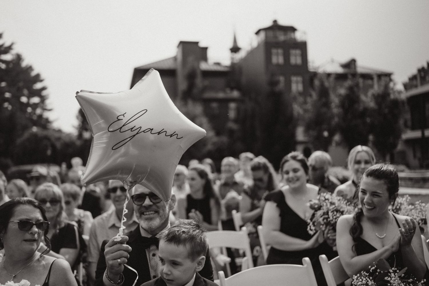 Black and white wedding ceremony photo of guests holding a star balloon with cursive writing outdoors.