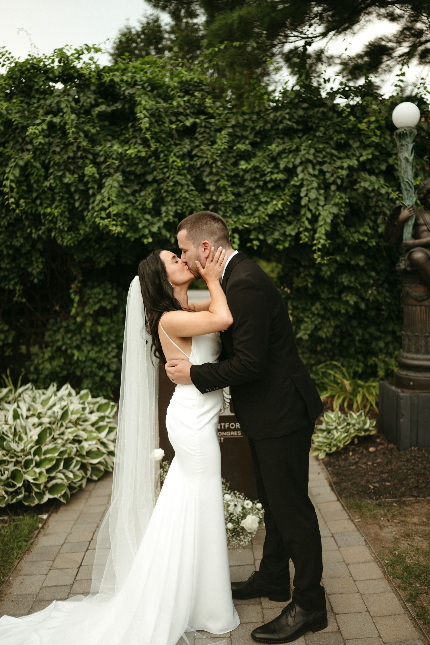 Bride and groom share a first kiss on a garden pathway surrounded by lush green hedges.