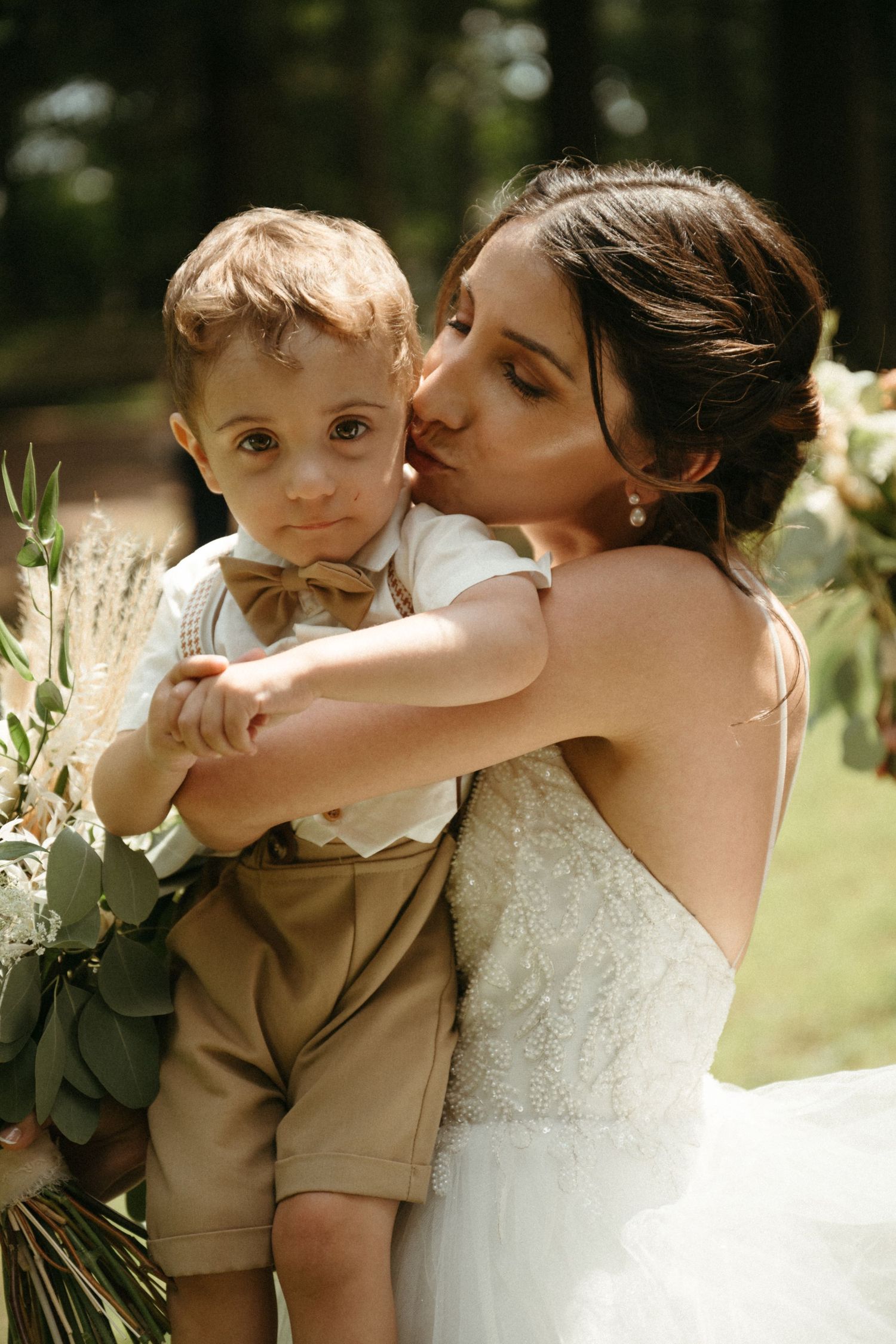 Bride in white lace gown tenderly kisses young ring bearer dressed in tan suspender outfit.