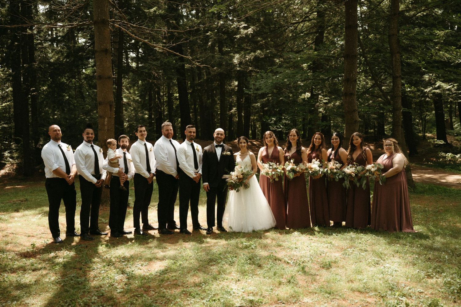 Wedding party posing together in a forest with bridesmaids in dusty rose dresses and groomsmen in black.