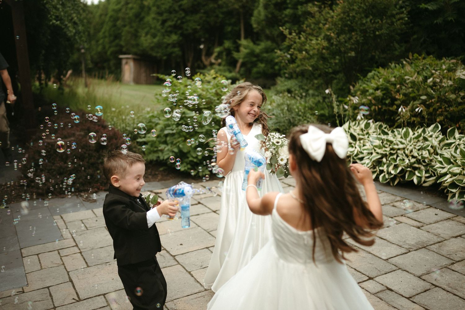 Three children in wedding attire play with bubble wands on a garden patio during a wedding.