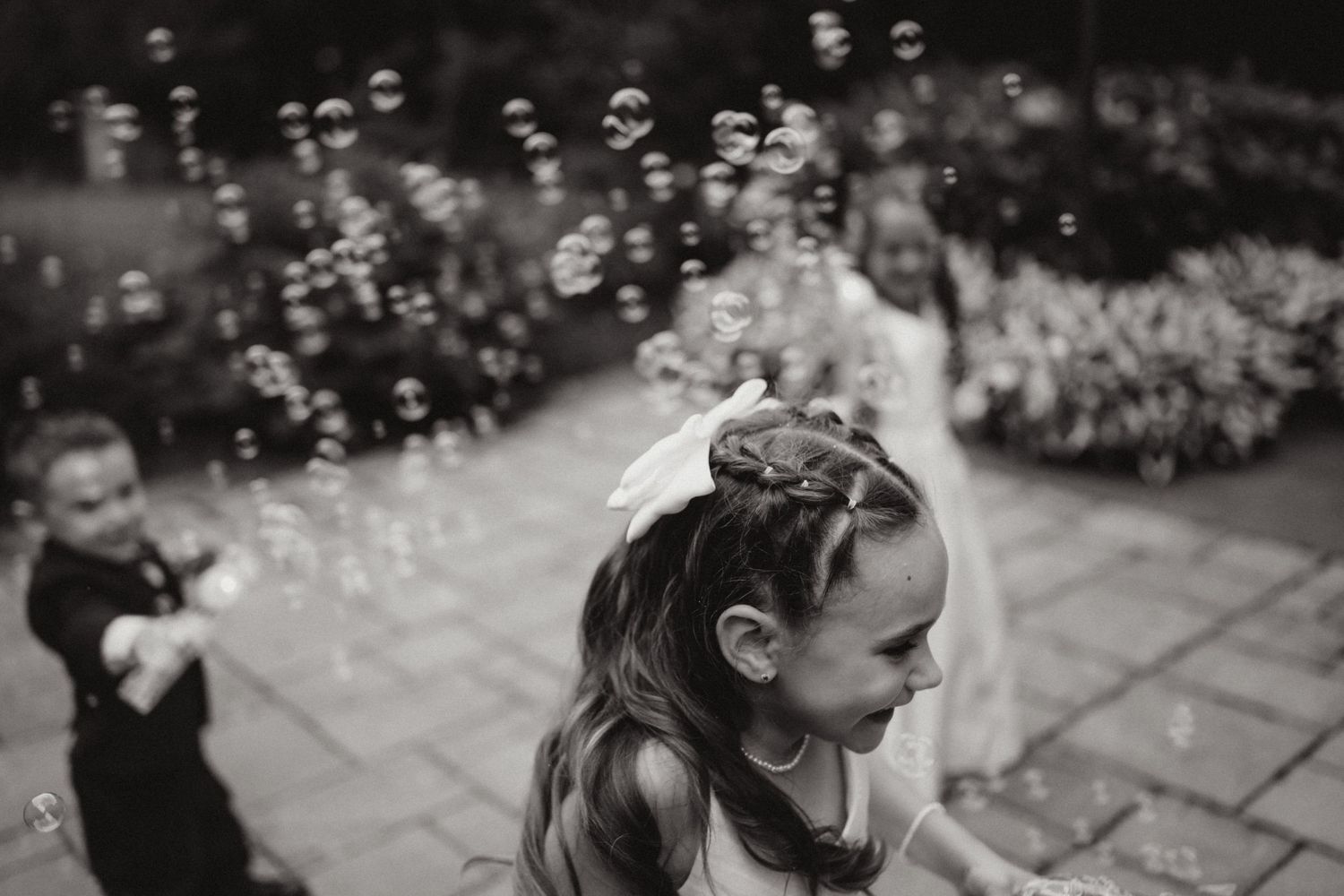 Black and white photo of flower girl playing with bubbles at an outdoor wedding celebration.