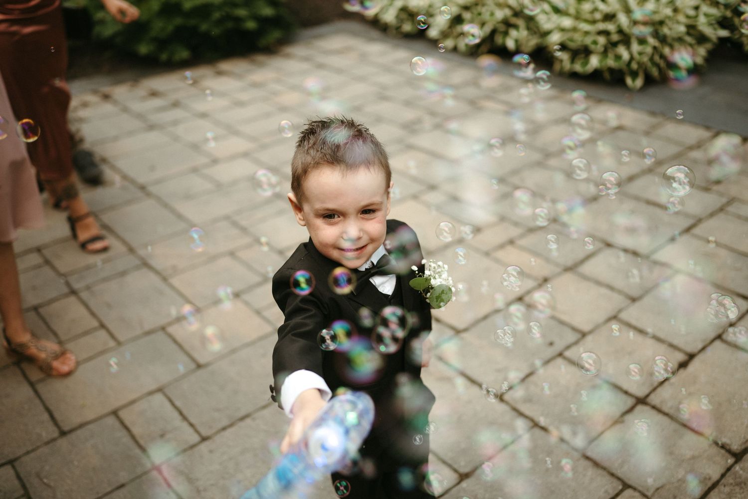 Smiling ring bearer in black suit blowing bubbles at a wedding on a brick patio.