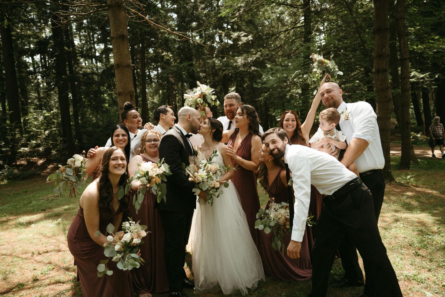 Wedding party celebrates with bride and groom in forest, bridesmaids in burgundy dresses holding bouquets.