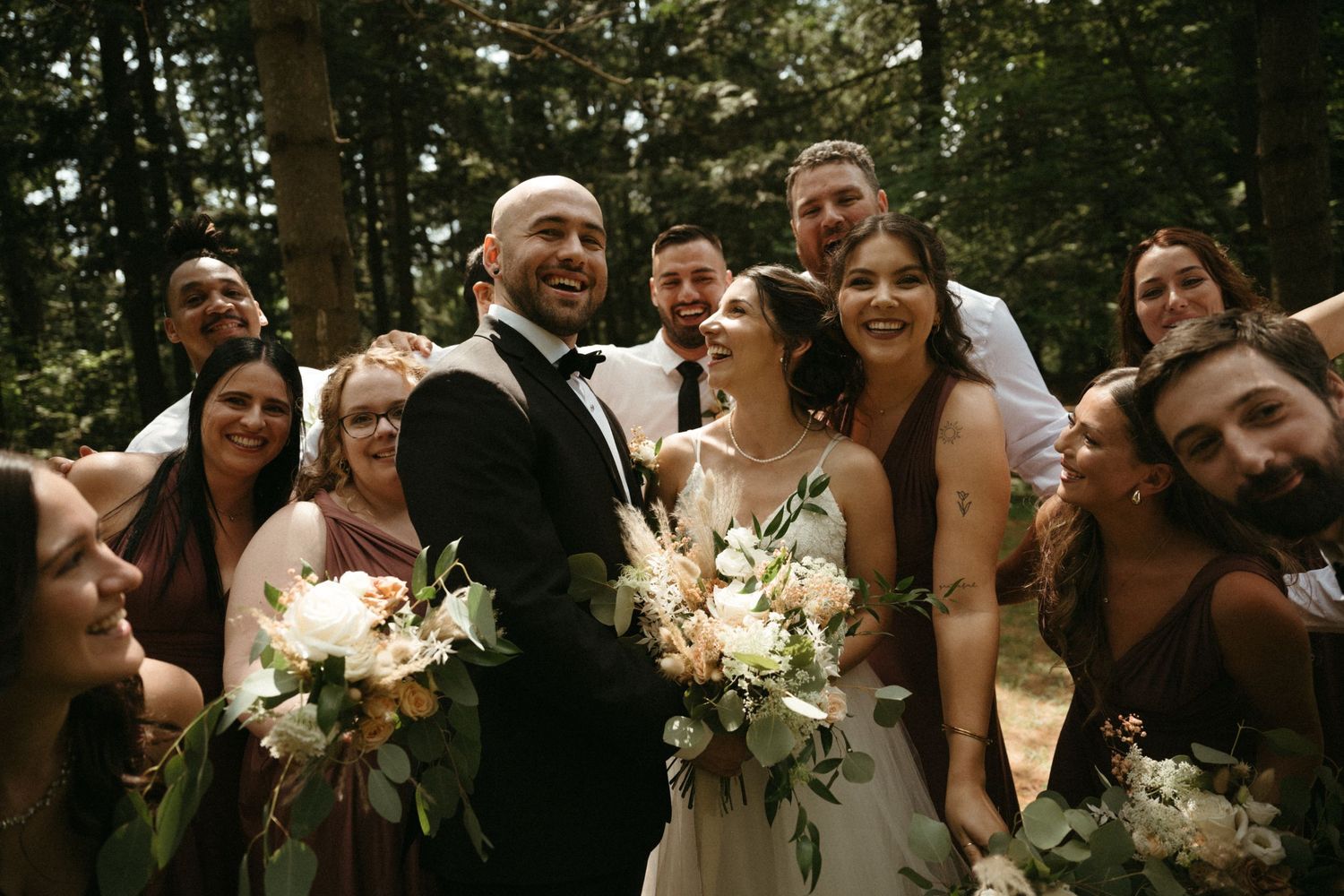 A happy bride and groom pose with their wedding party surrounded by tall trees in a forest.