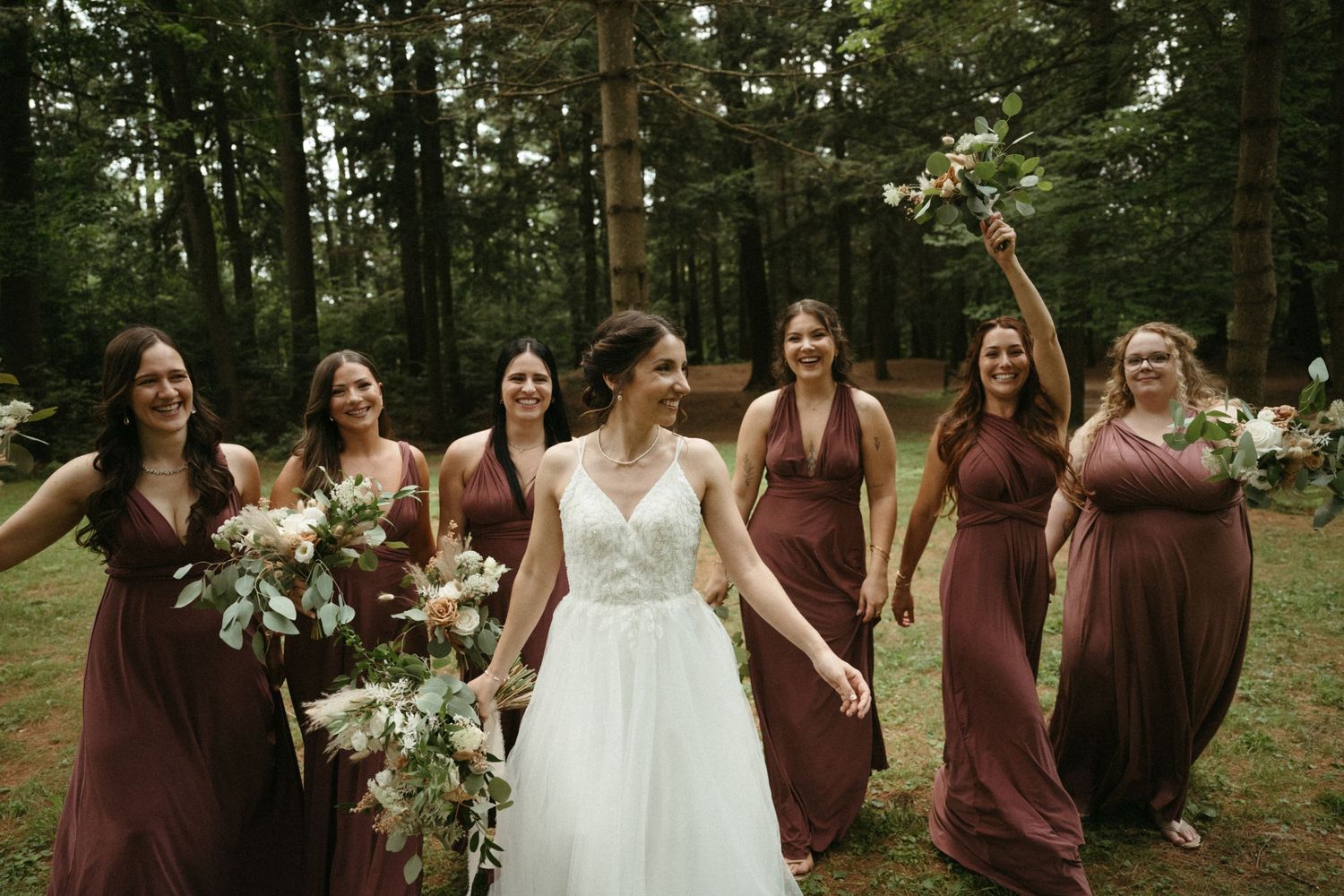 Bride in white lace gown walks joyfully with six bridesmaids in burgundy dresses through a forest wedding venue.