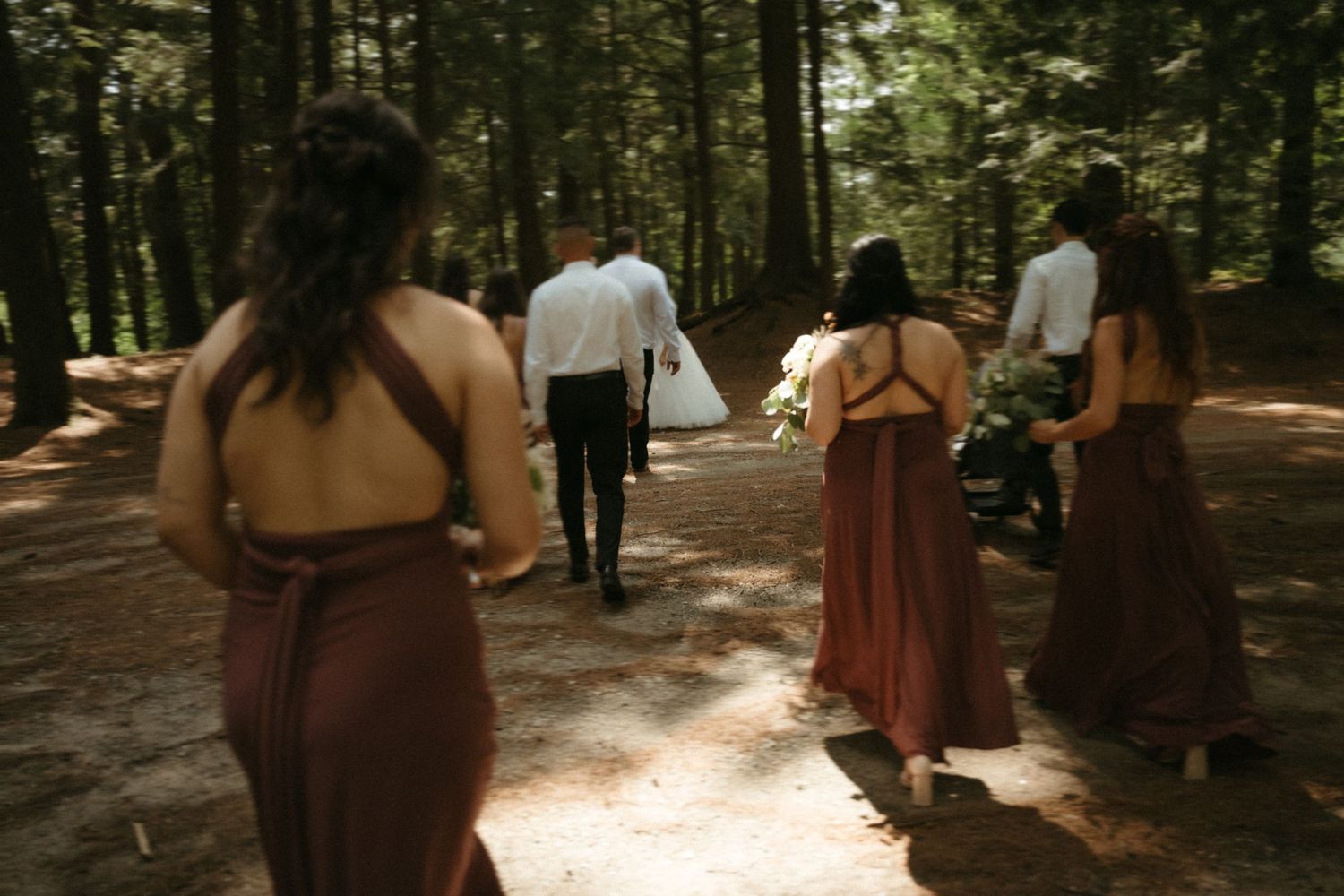 Bridesmaids in rust dresses walk with wedding party along a forest path toward ceremony arch.