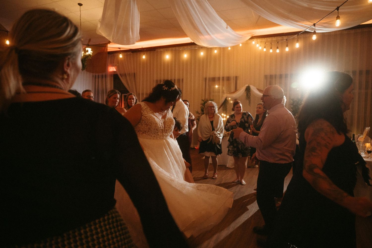 Bride in white gown dancing with guests at elegant wedding reception with warm string lights.