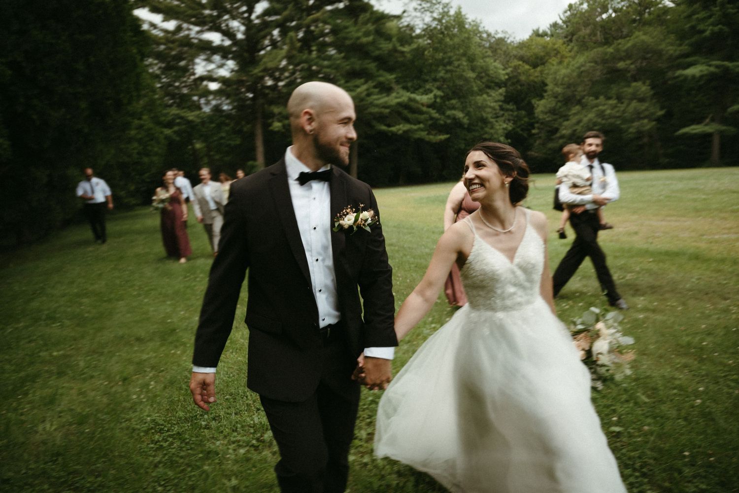 Bride and groom holding hands walking on green lawn at outdoor wedding reception smiling joyfully.