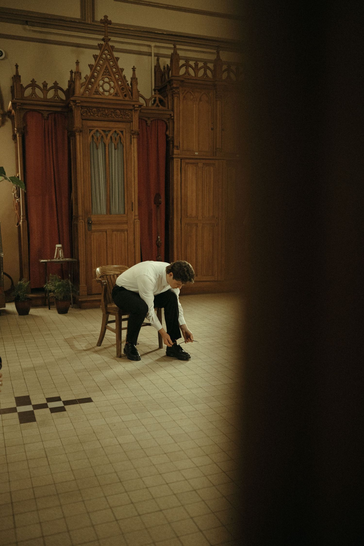 Groom tying his shoes beside ornate wooden Gothic cabinets on a tiled floor before his wedding ceremony.