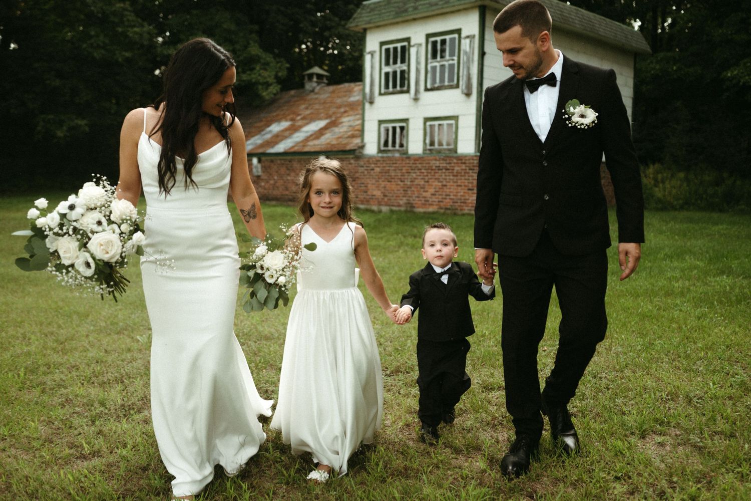 Bride and groom in formal attire walk with flower girl and ring bearer near rustic venue.