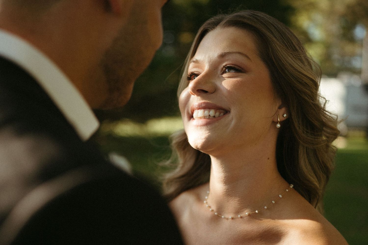 A smiling woman wearing a white dress and necklace, standing outdoors in a garden setting.