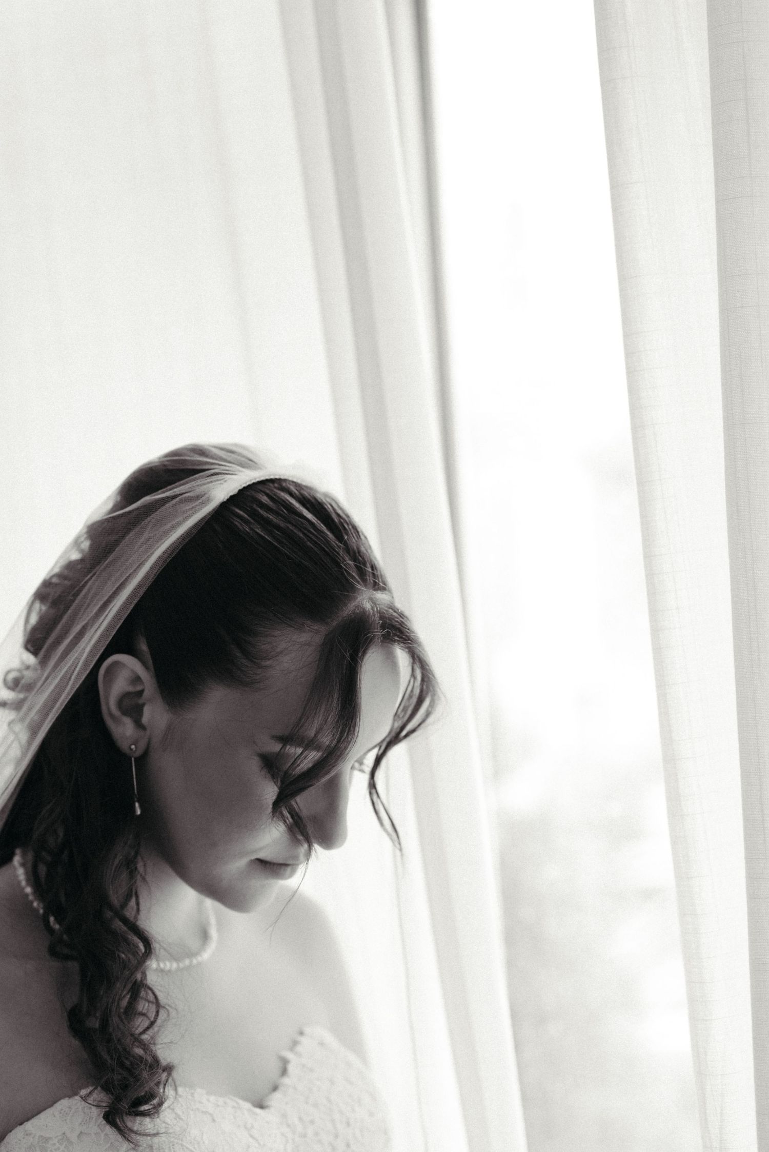 Black and white portrait of bride in veil gazing down near sheer curtained window.