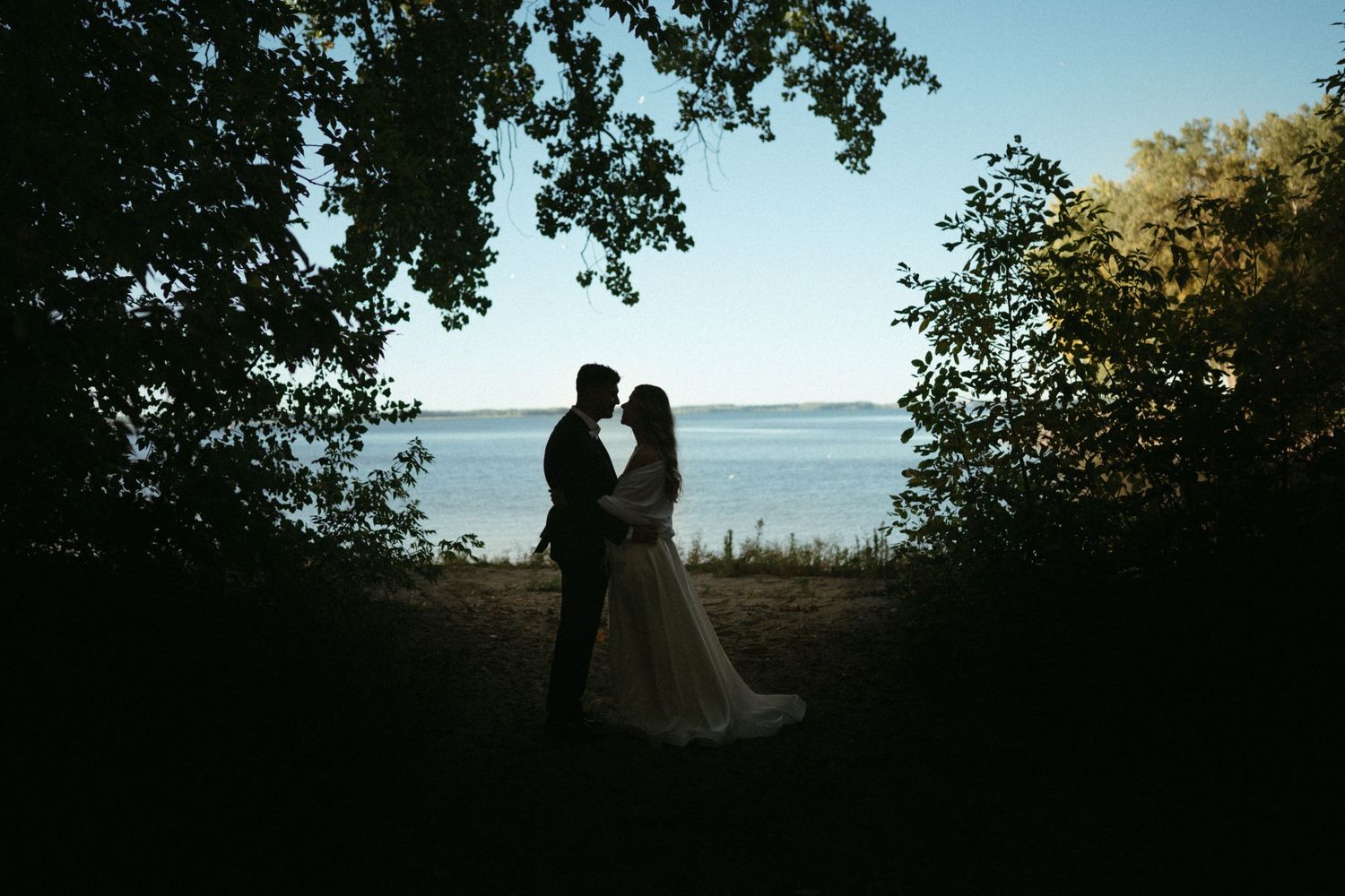 A silhouetted couple embracing in a scenic outdoor setting with trees and a body of water in the background.