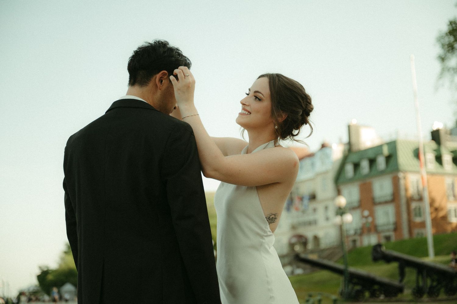 A smiling bride in a white dress tenderly touches her groom's face outdoors near a historic building at dusk.