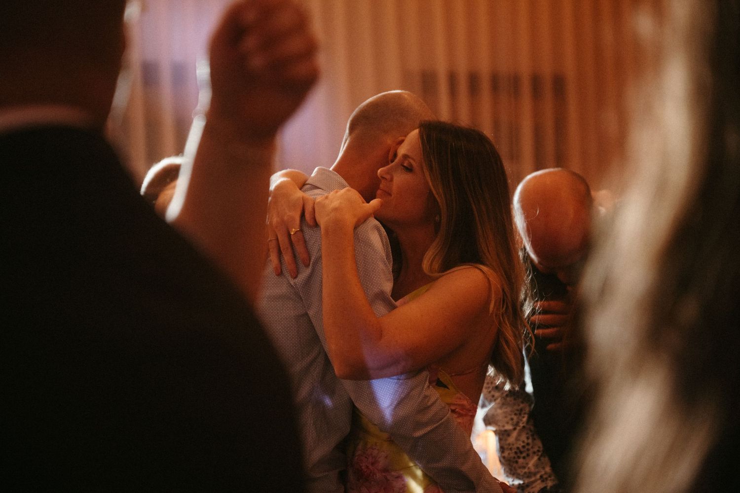 Couple sharing a romantic kiss on the dance floor surrounded by guests at a dimly lit wedding reception.