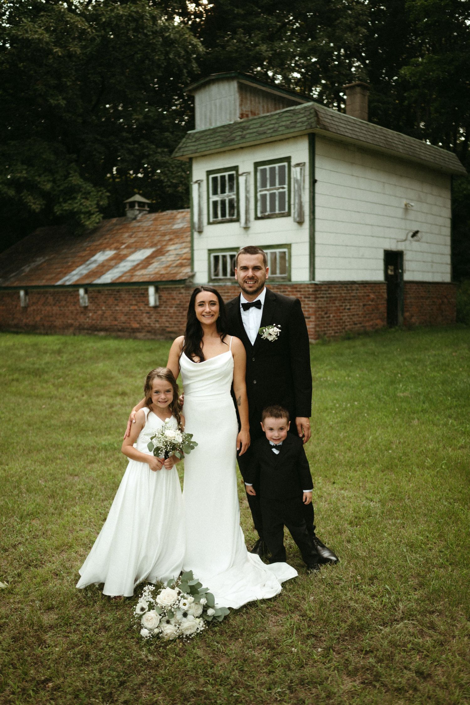 Wedding couple poses with flower girl and ring bearer in front of a charming white and brick building.