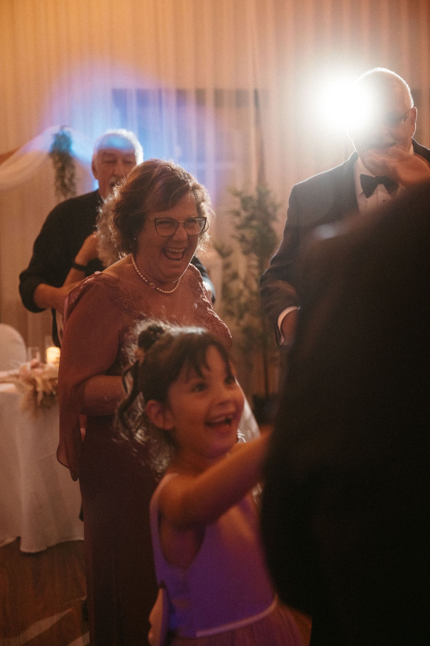 A grandmother laughs joyfully while dancing with a young flower girl at a wedding reception.