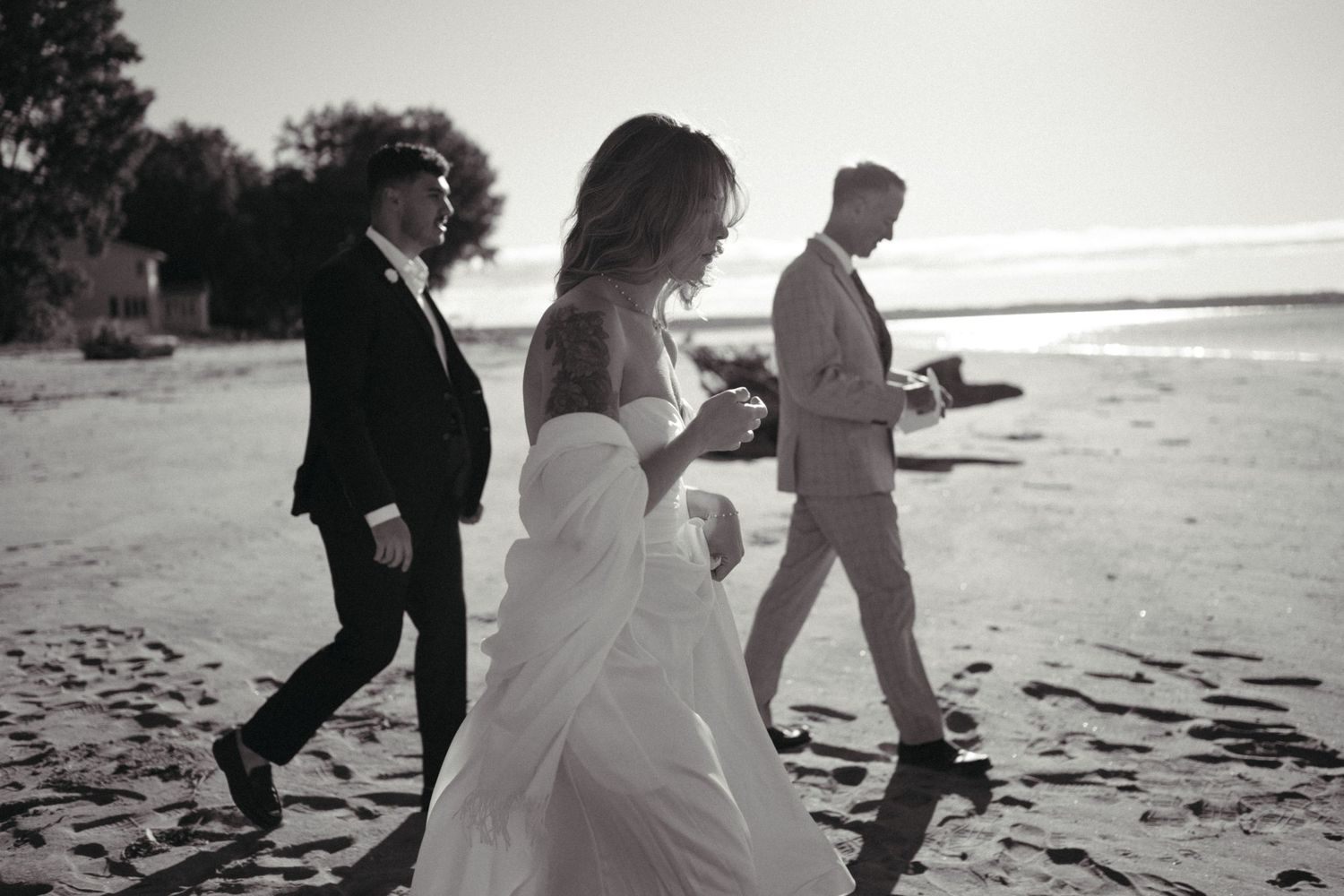 A bride and groom walking on the beach with a third person, captured in a black and white photograph.