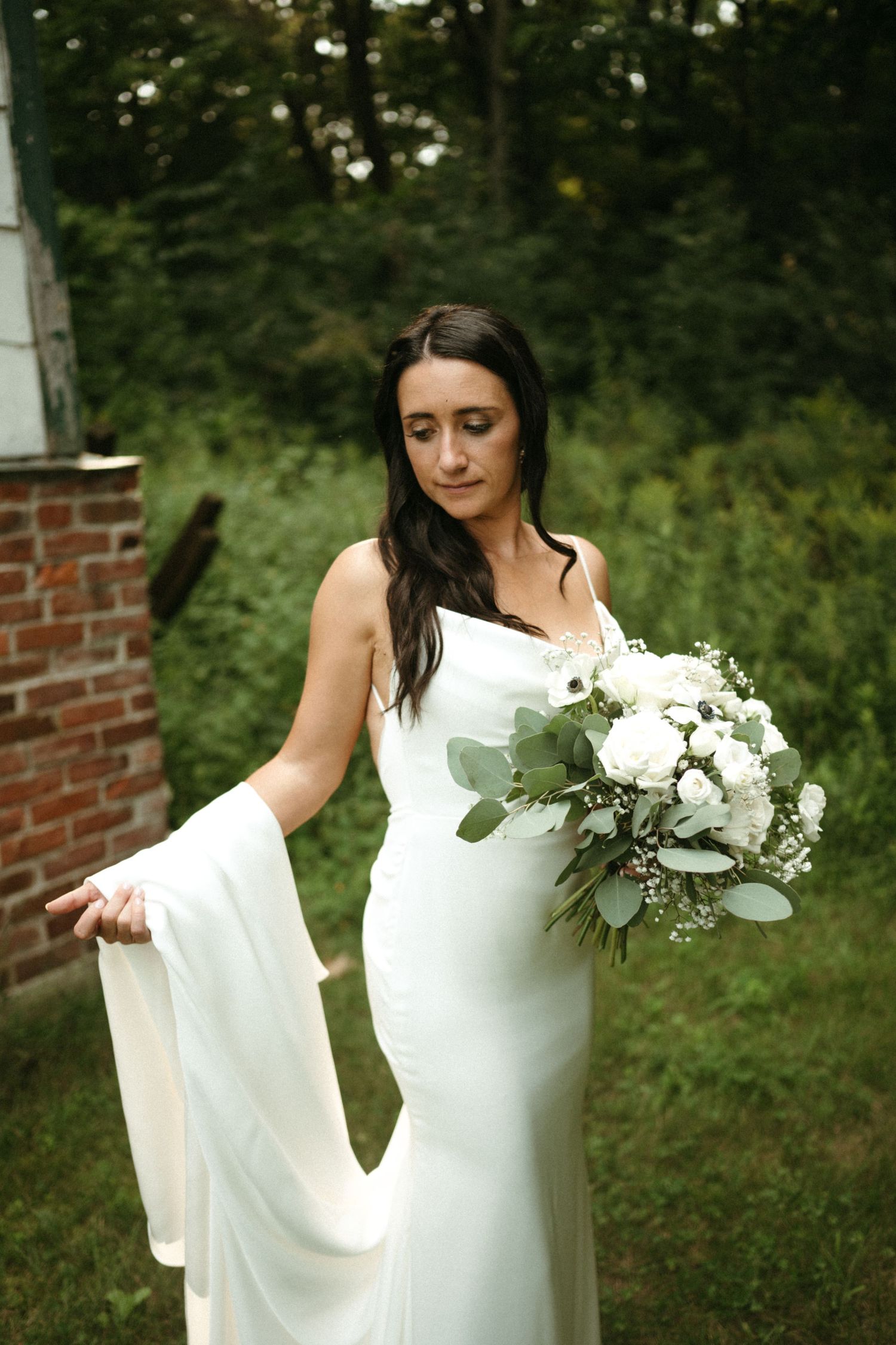 Bride in sleek white gown holding white floral bouquet with greenery outdoors near brick wall.