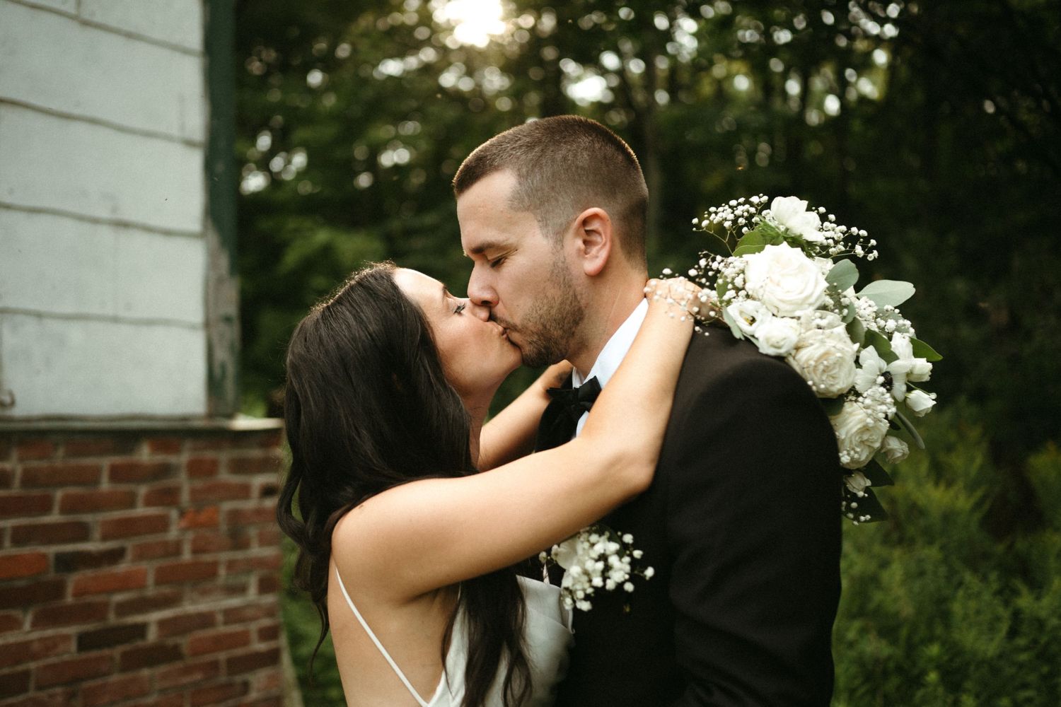 Bride and groom sharing a kiss outdoors, bride holding white floral bouquet near rustic brick building.