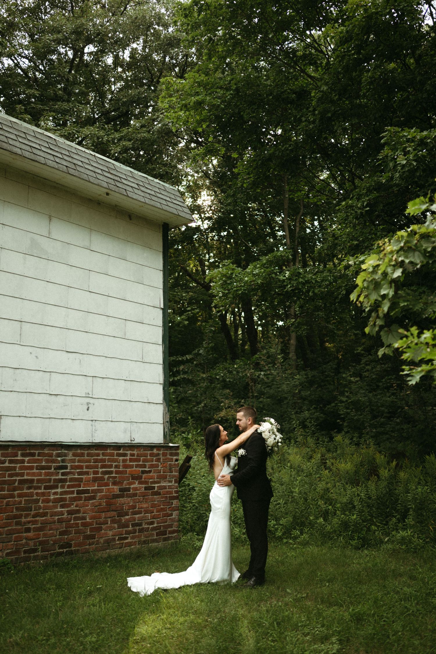 Bride and groom embracing near a rustic brick building surrounded by lush green trees on their wedding day.