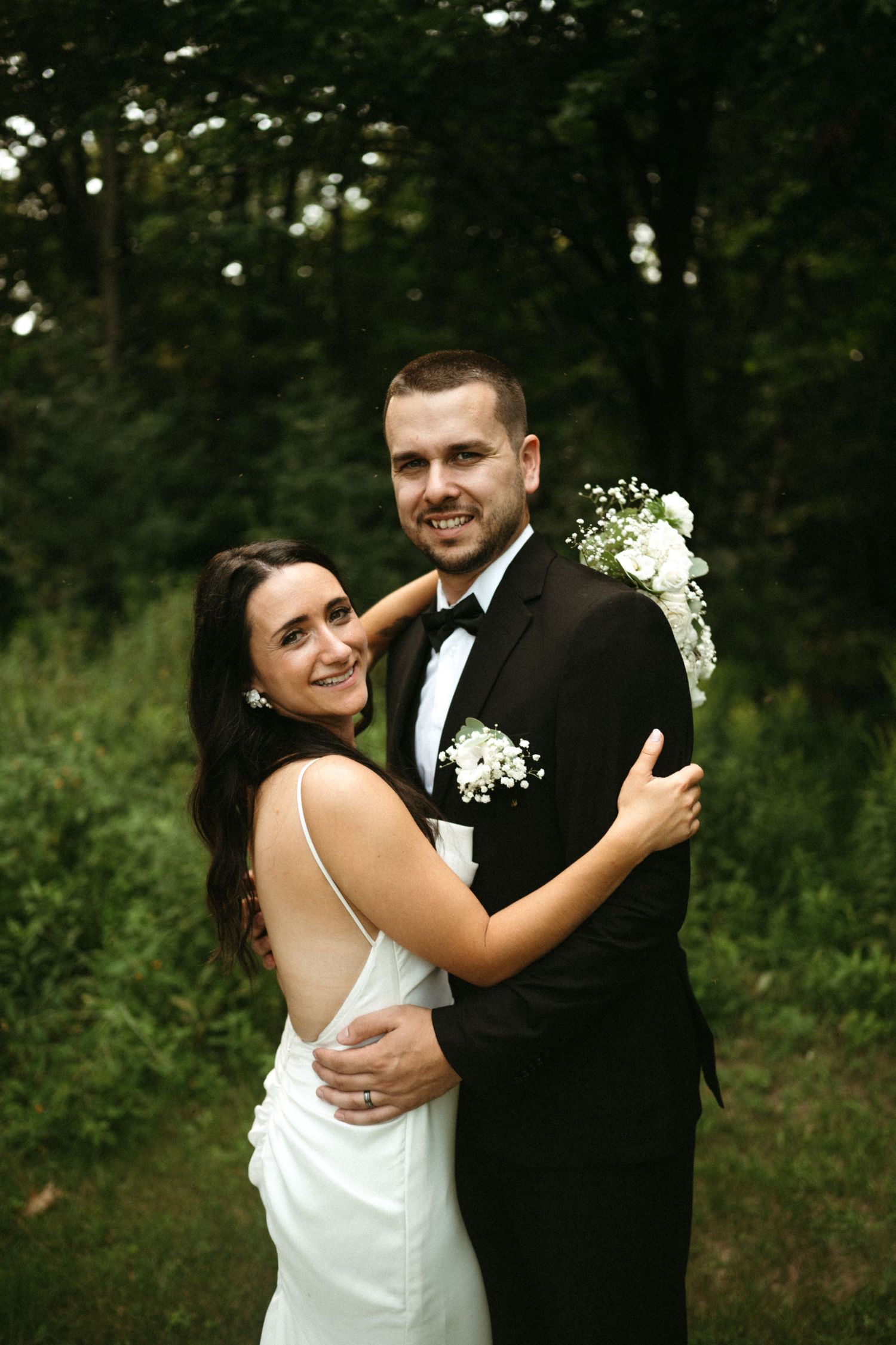 Bride in white gown and groom in black tuxedo embrace in a lush green forest setting.