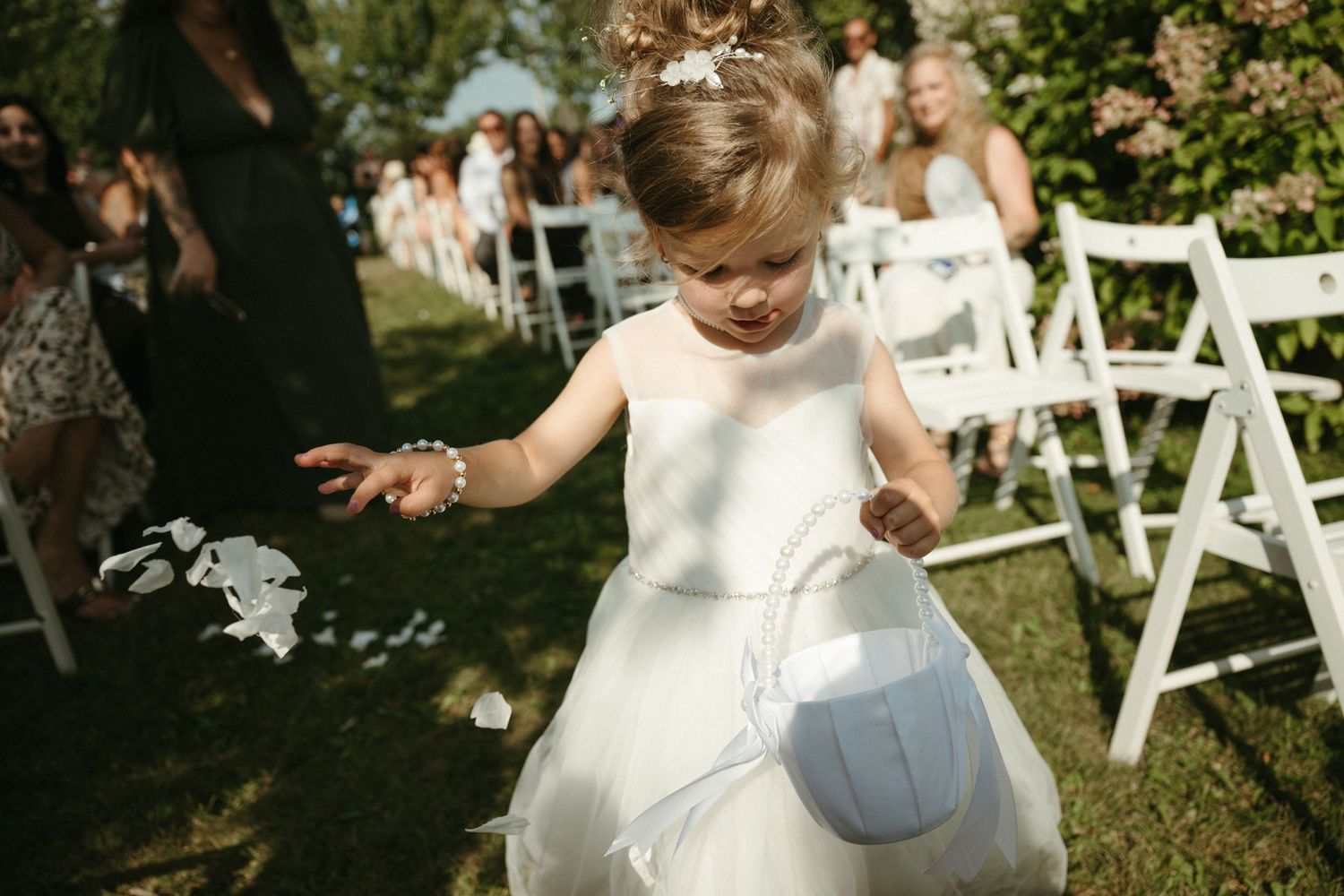 A young flower girl in a white dress scatters petals down the aisle at an outdoor wedding ceremony.