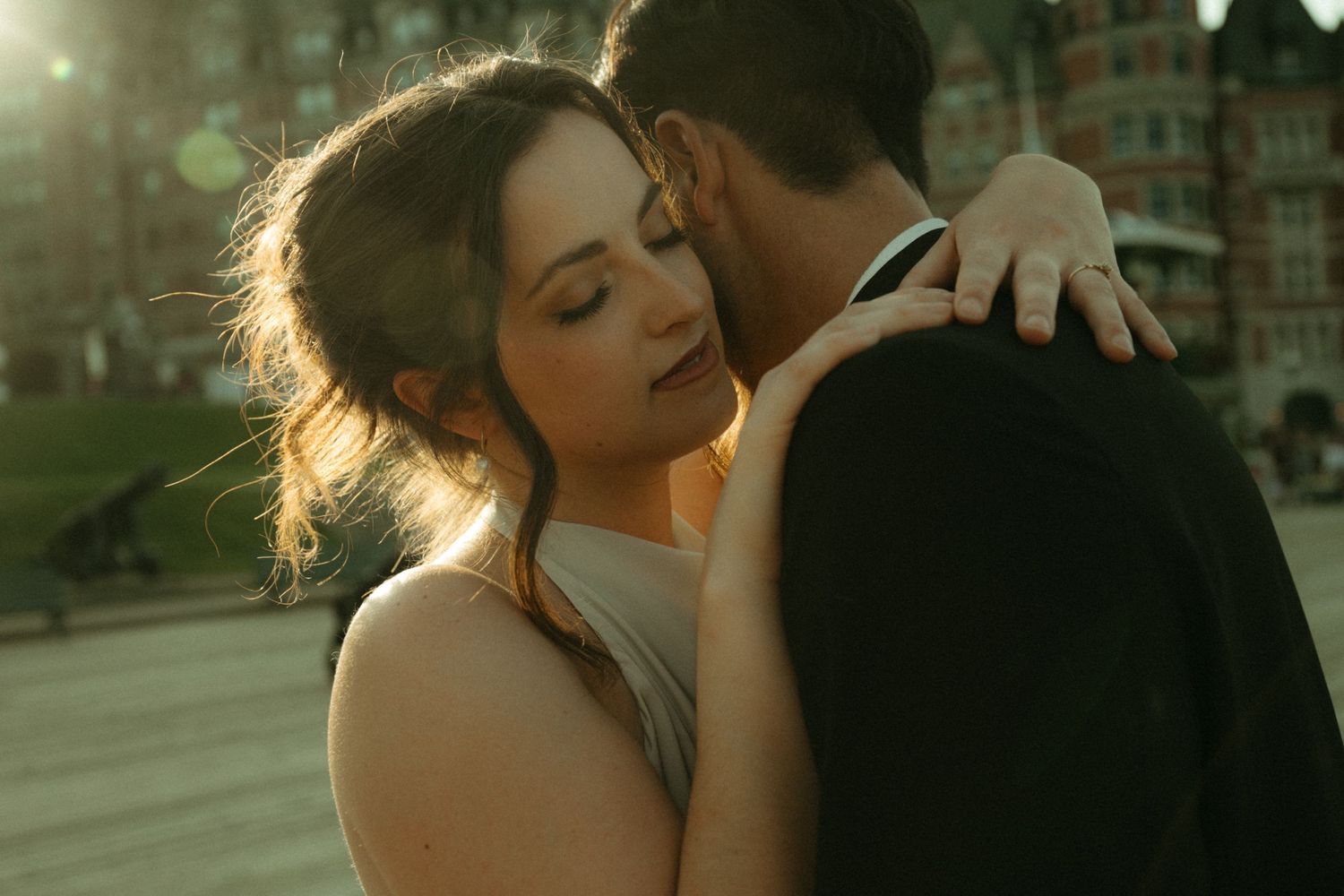 A couple shares a romantic embrace at golden hour, with the bride resting her head near the groom's shoulder outdoors.
