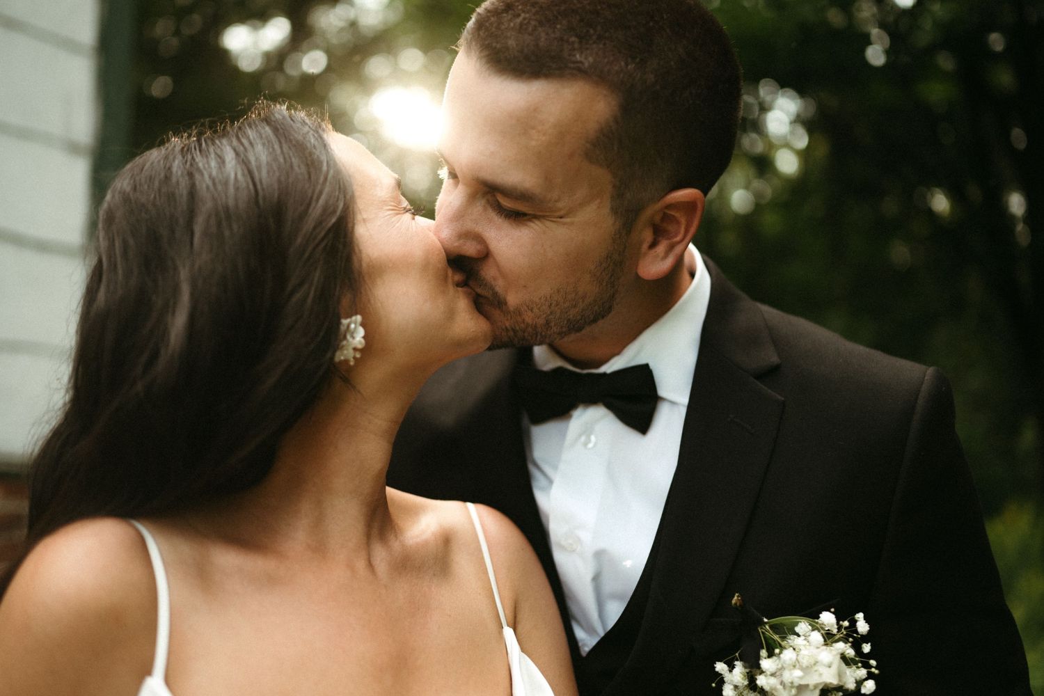 Bride and groom sharing a romantic kiss outdoors with warm sunlight glowing behind them.