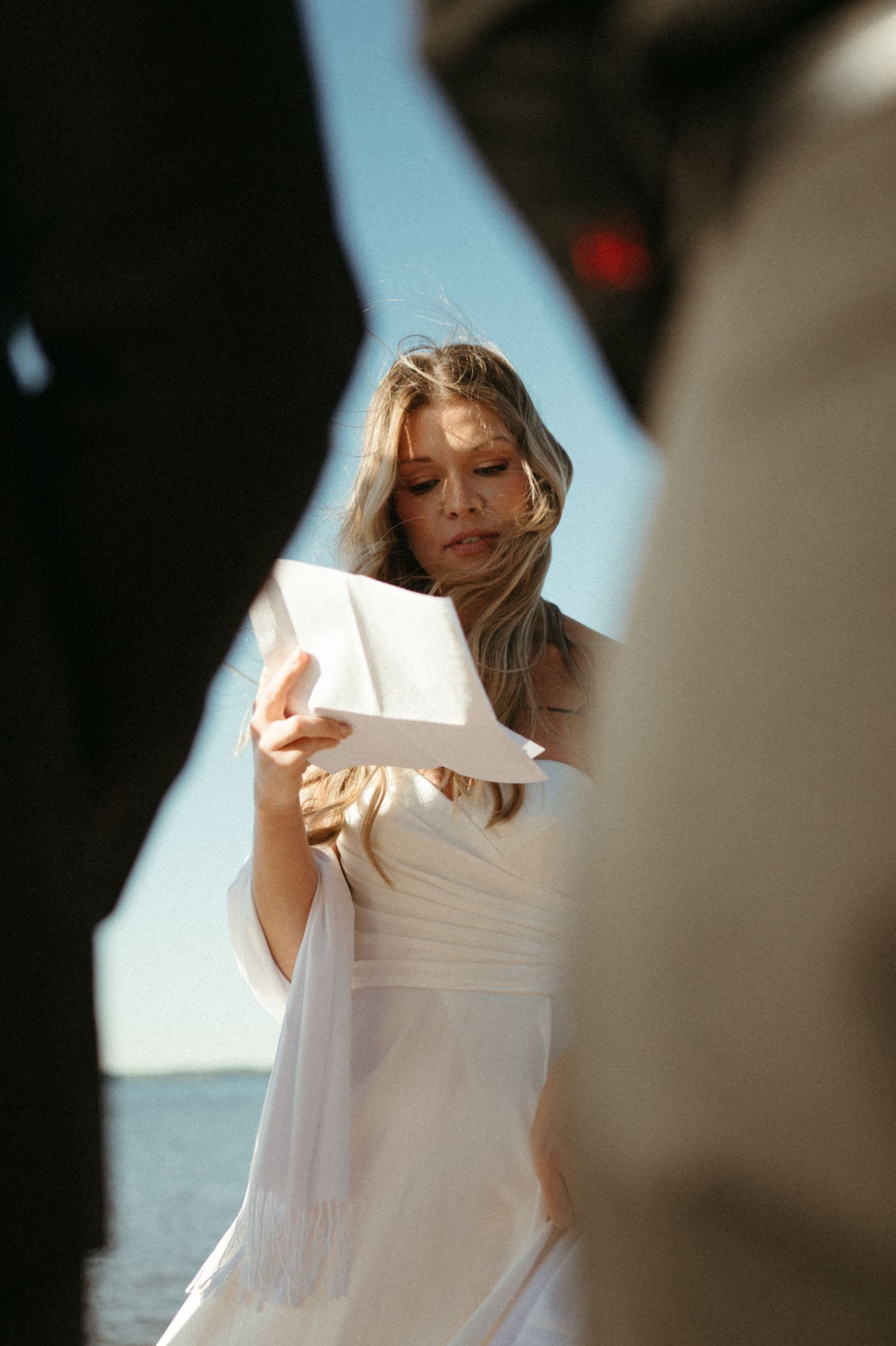 A bride holds a book and stands in a sunlit outdoor environment.