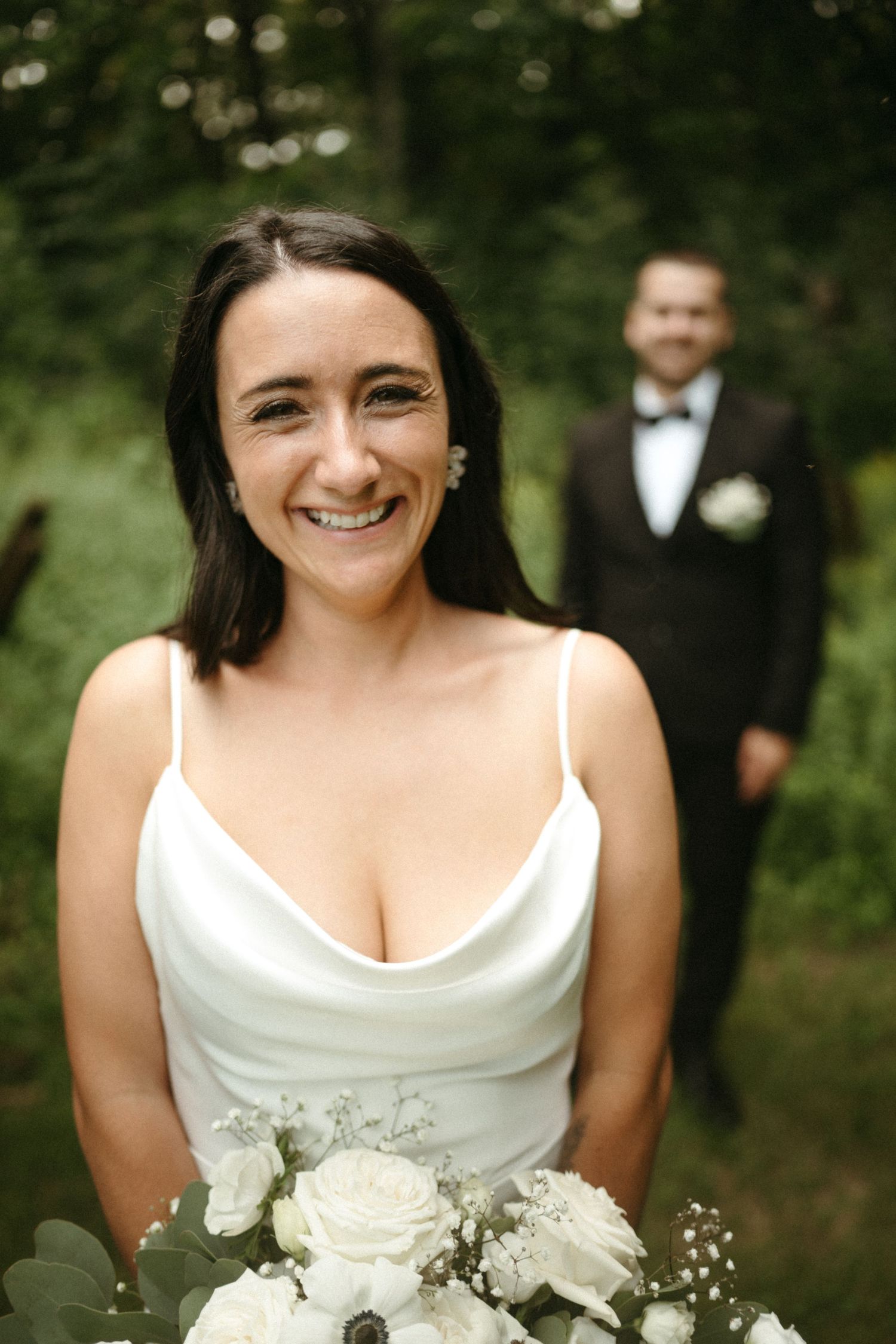 Smiling bride in white satin gown holding white rose bouquet with groom in tuxedo standing in green forest background.