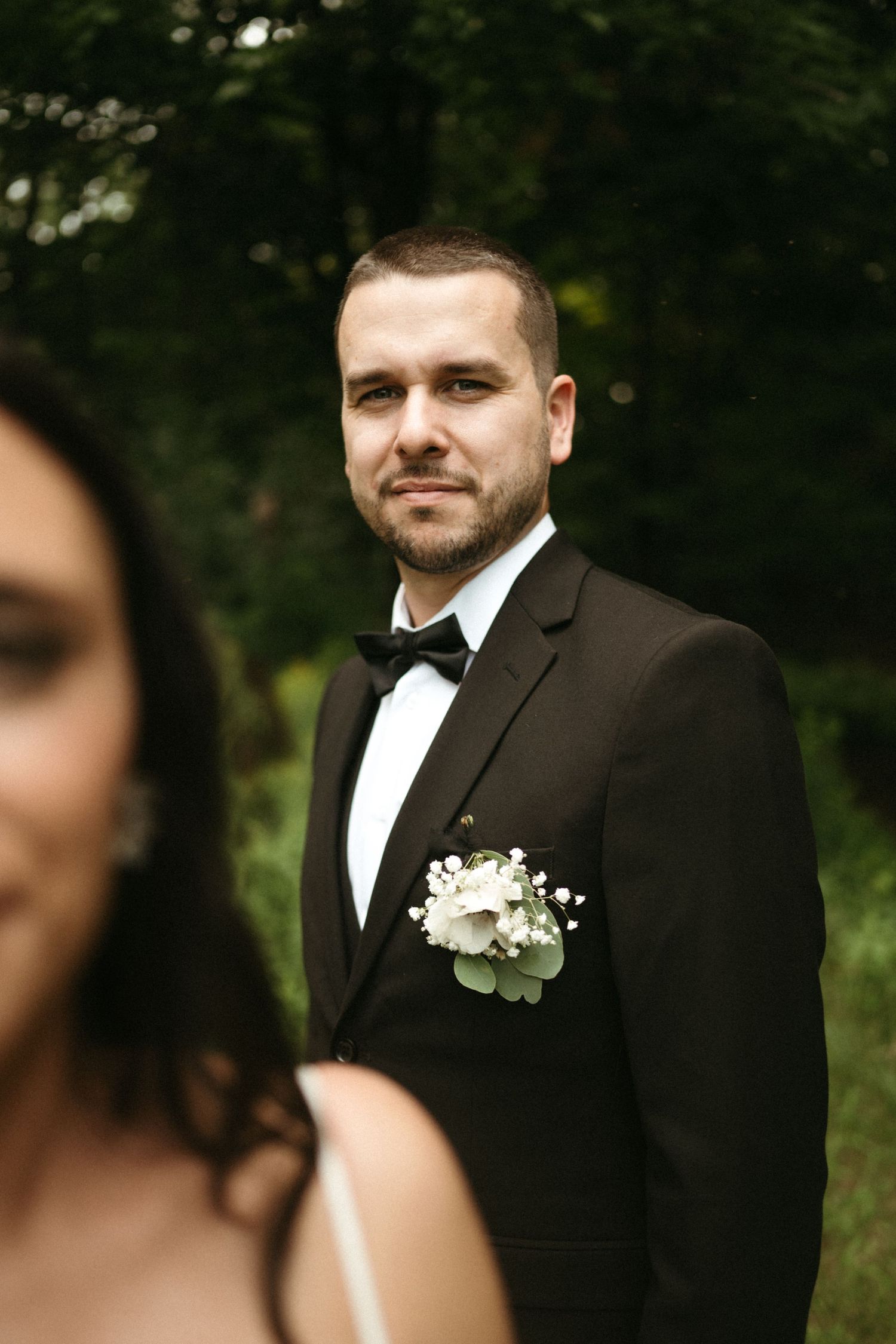 Groom in black tuxedo with white boutonniere standing outdoors with lush green trees in background.