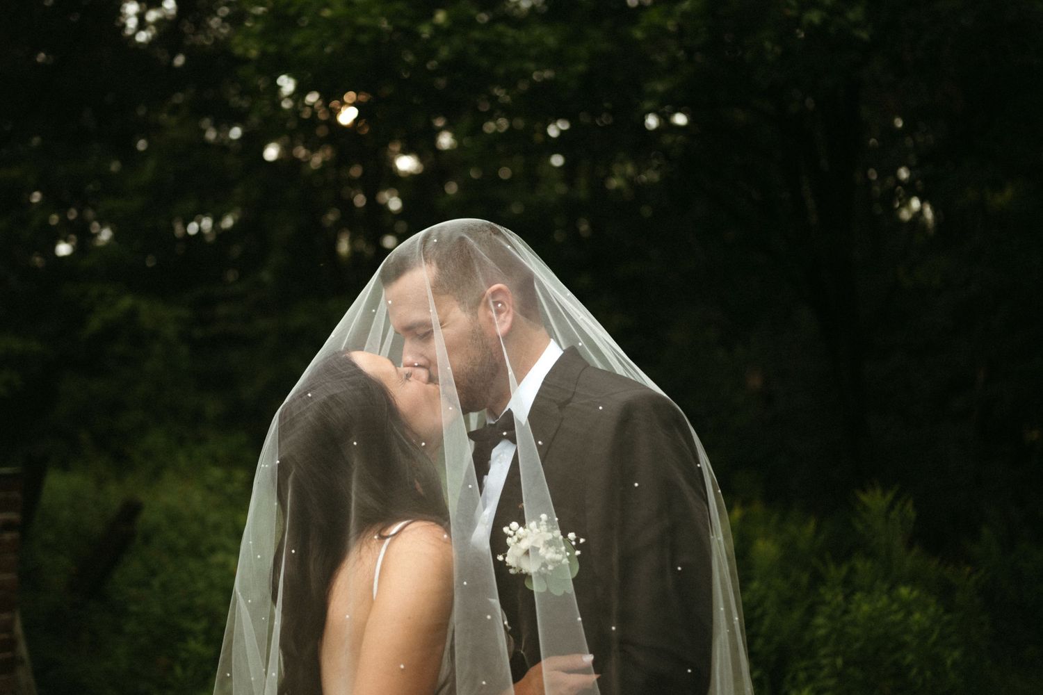 Bride and groom share romantic kiss under sheer veil outdoors with dark green trees behind them.
