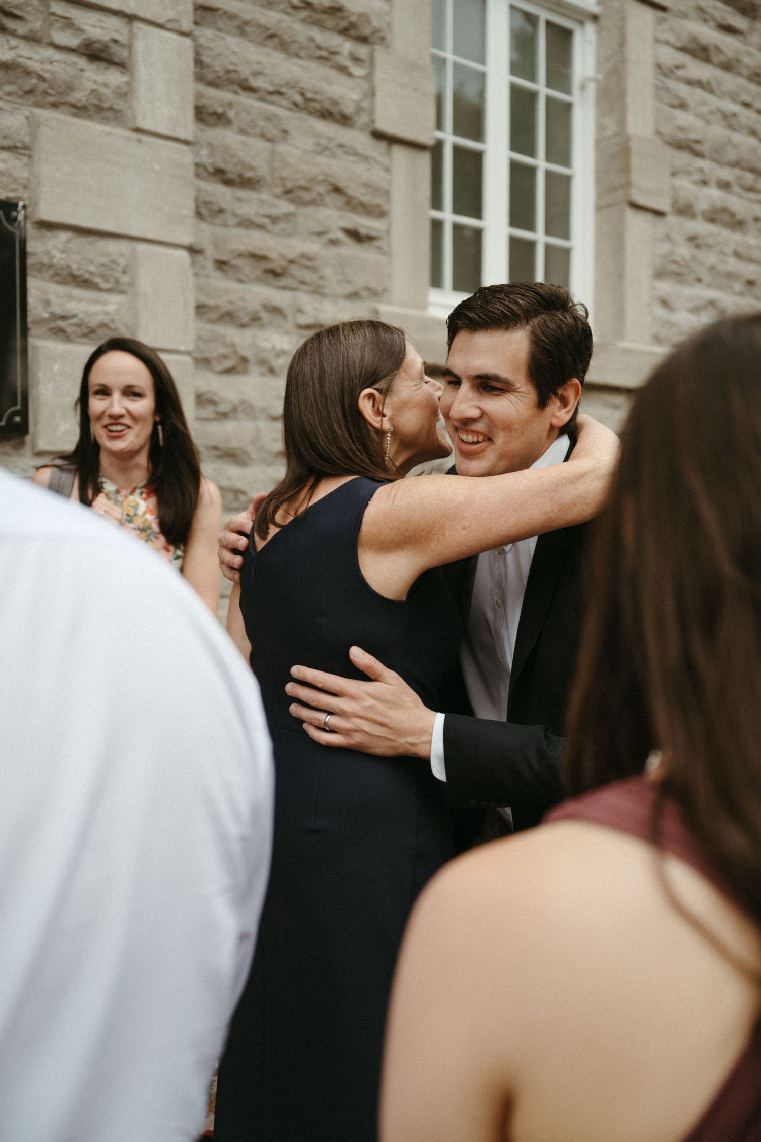 Wedding guest in black dress shares warm embrace with smiling young man outside venue.