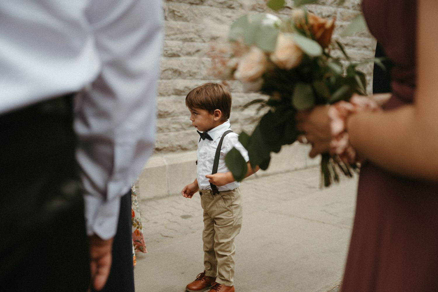 Adorable young ring bearer in khaki pants and suspenders standing during outdoor wedding ceremony holding bouquet.