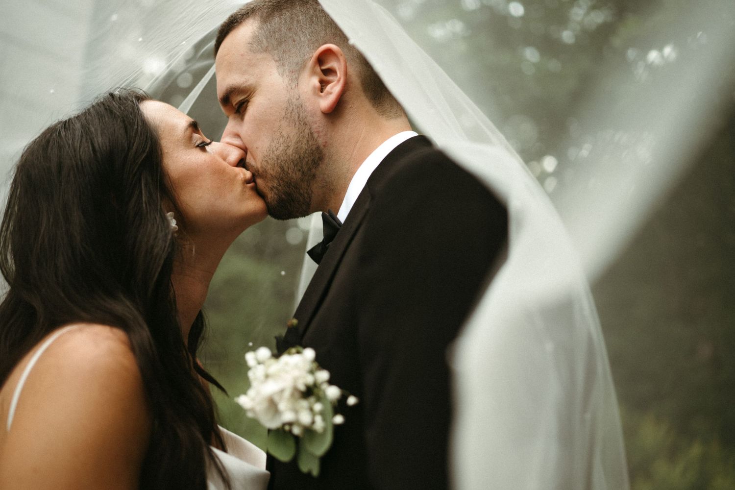 Bride and groom share a romantic kiss under a flowing white veil in a lush outdoor setting.