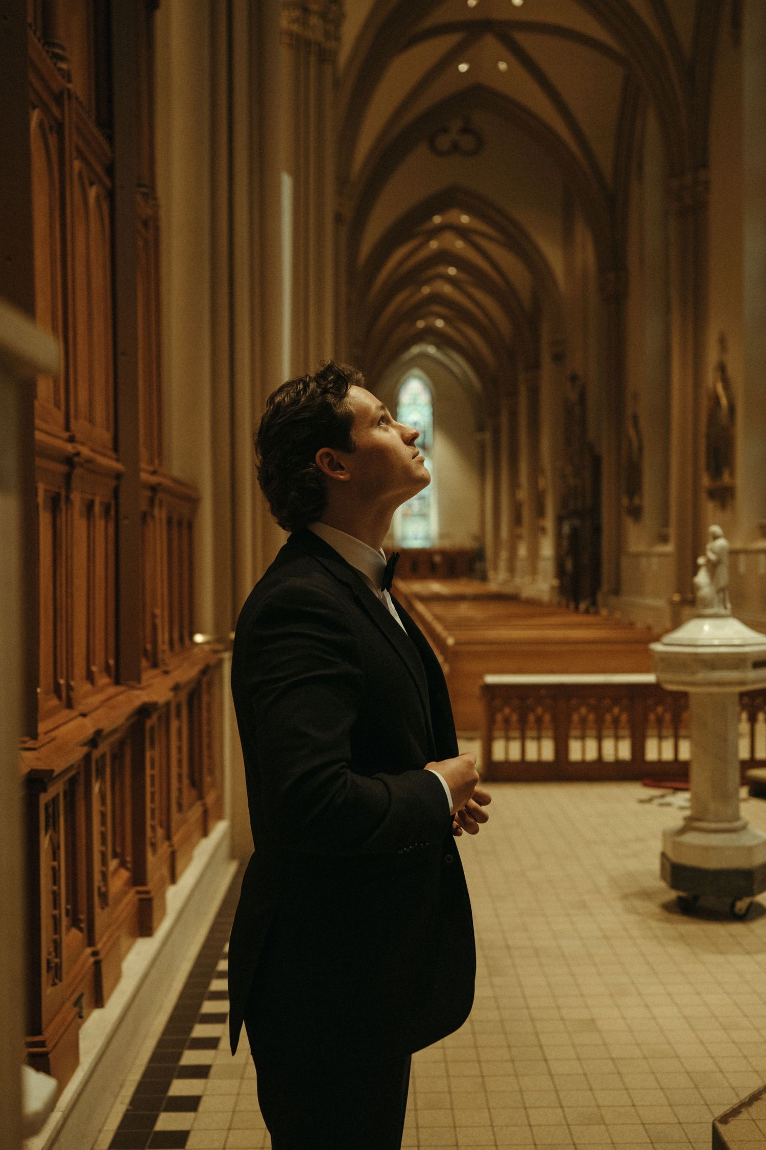 A groom in a black tuxedo gazes upward in awe inside a stunning Gothic cathedral with arched ceilings.