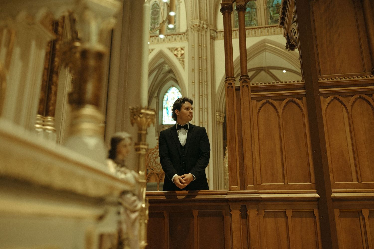Groom in black tuxedo sitting quietly in ornate Gothic cathedral pew before wedding ceremony begins.