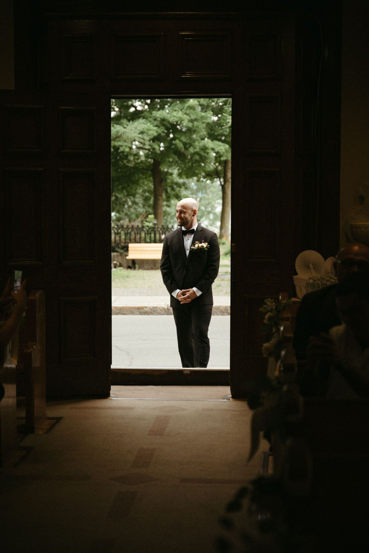 Groom in black tuxedo standing in a dramatic doorway of a dark church before the ceremony.