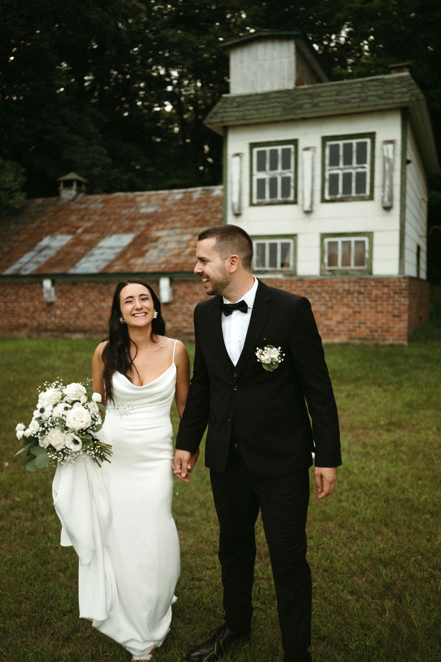 Bride and groom holding hands walking outdoors in front of a white farmhouse, bride in white gown with bouquet.