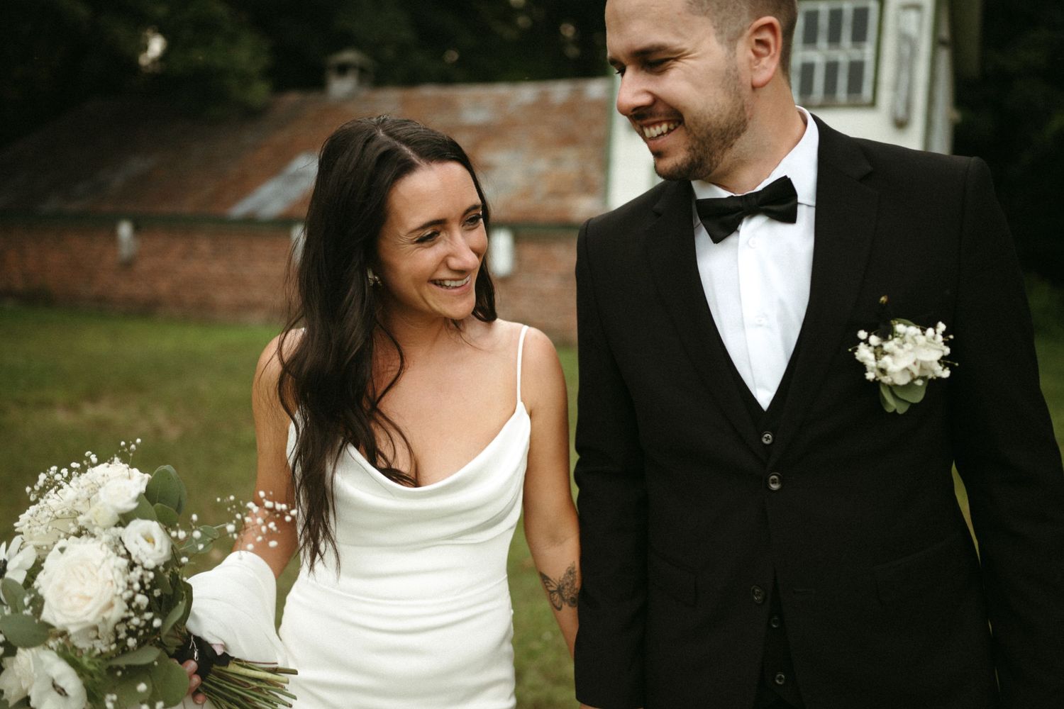 Smiling bride holding white bouquet and groom in black tuxedo walk together at outdoor wedding.