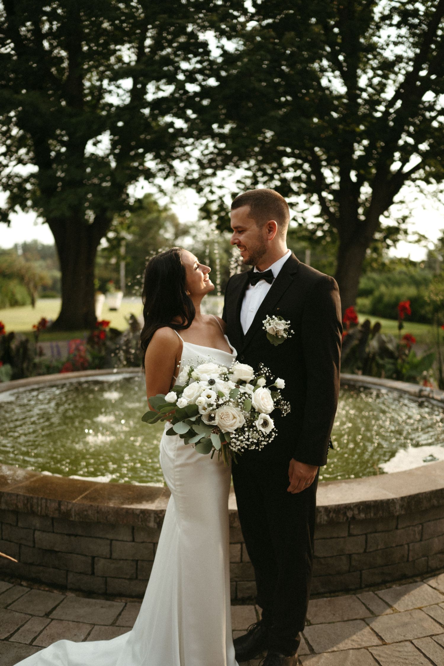 Bride and groom gazing at each other near an elegant garden fountain, bride holding a white floral bouquet.