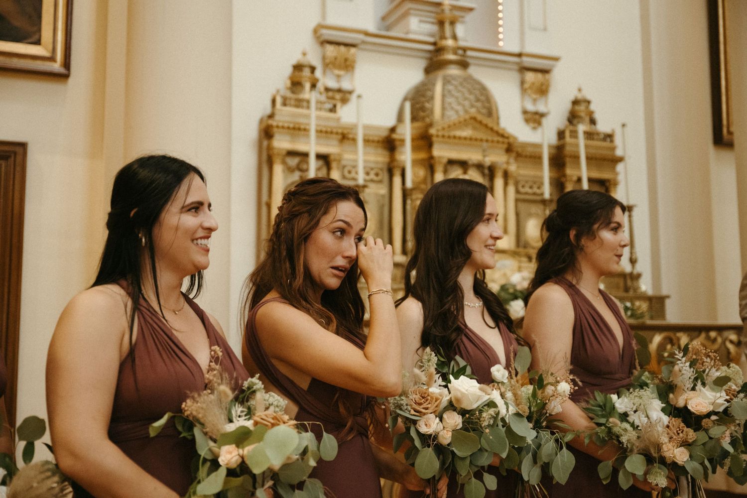 Bridesmaids in mauve dresses holding floral bouquets, one wiping tears, inside ornate church.