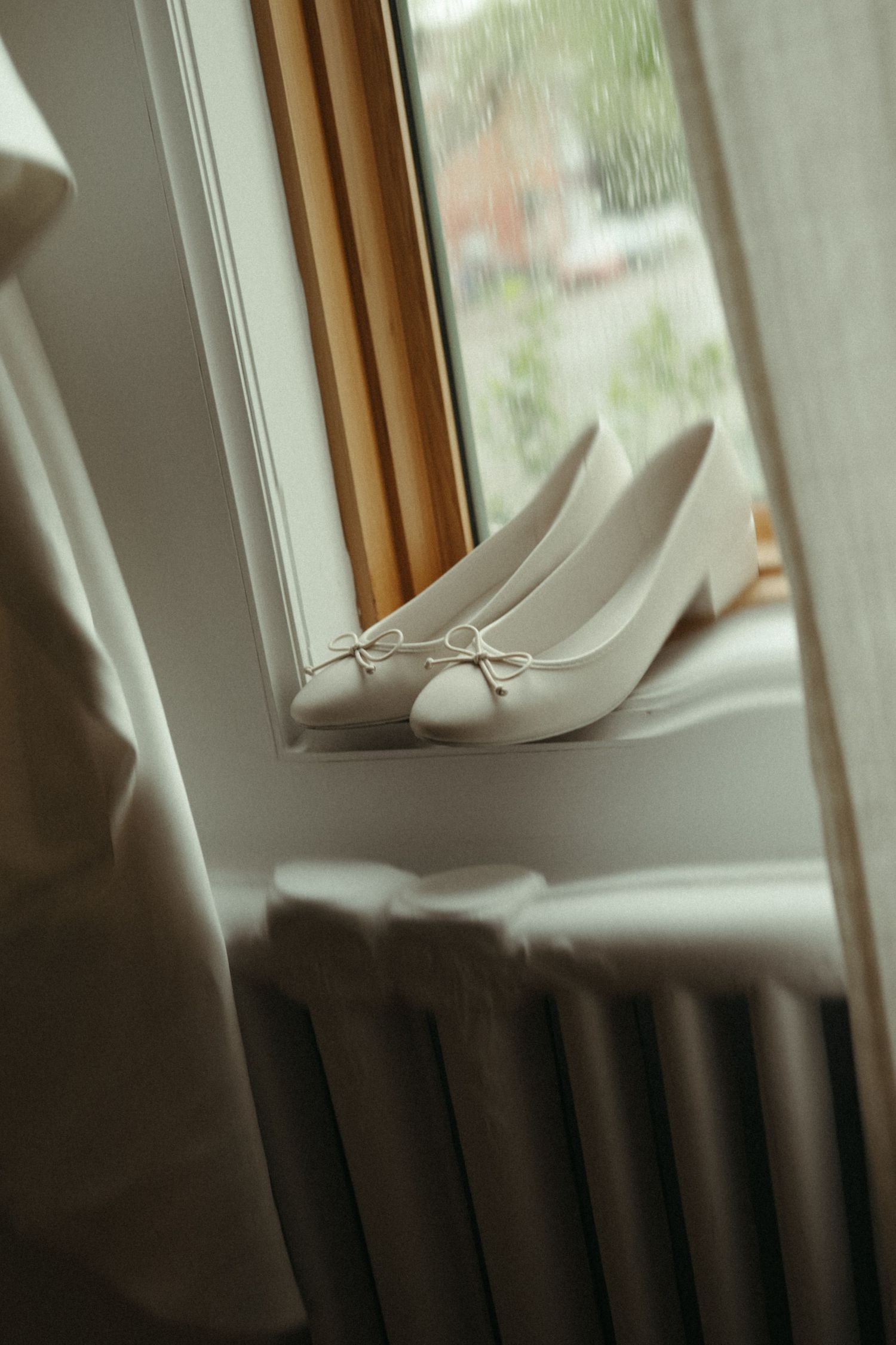White bridal ballet flats with delicate bow detail displayed on windowsill with raindrops visible outside window.