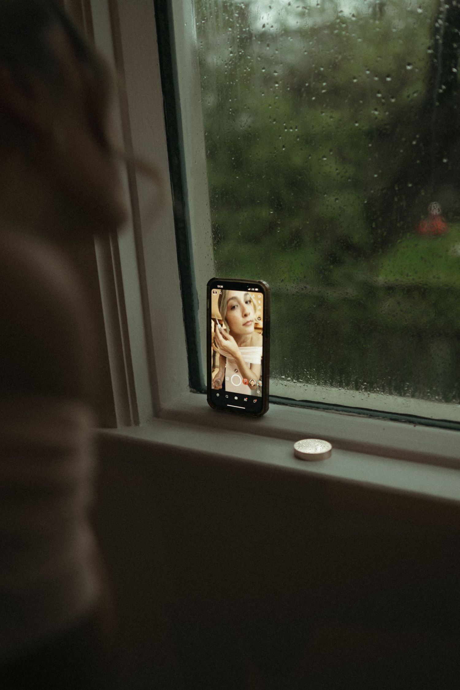 Bride's photo displayed on smartphone propped on rainy window sill before wedding day.